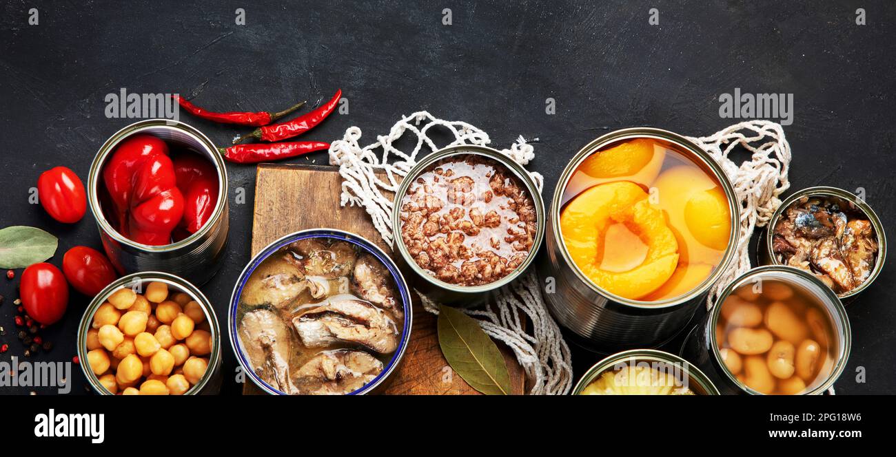 Various canned vegetables, meat, fish and fruits in tin cans. On a dark background. Top view. Panorama with copy space. Stock Photo