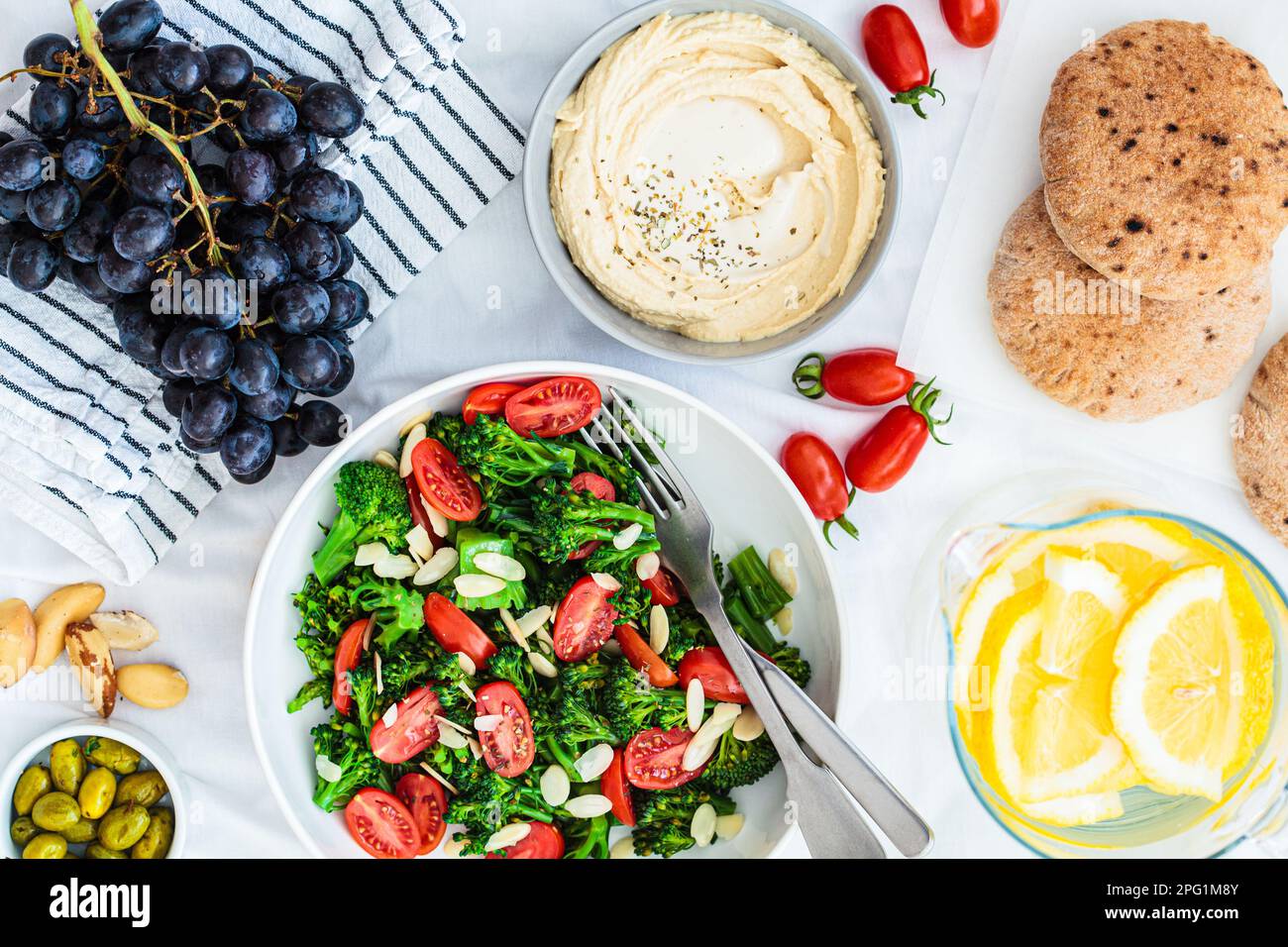 Vegan picnic food: hummus, lemonade, broccoli salad and fruits on a white blanket on the grass, top view. Stock Photo