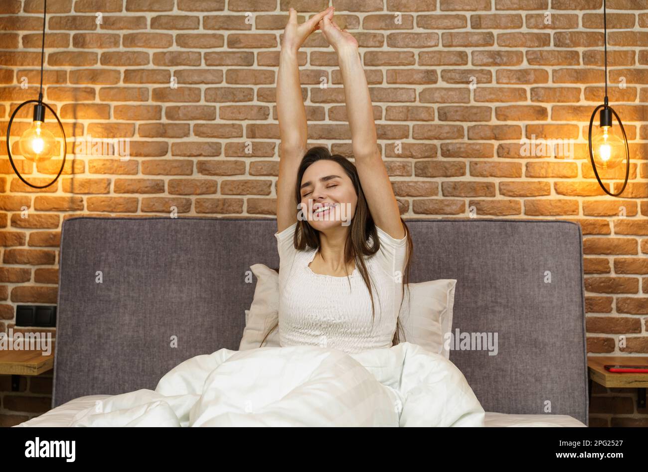 Beautiful woman sits on the bed, stretches her arms up after sleeping ...