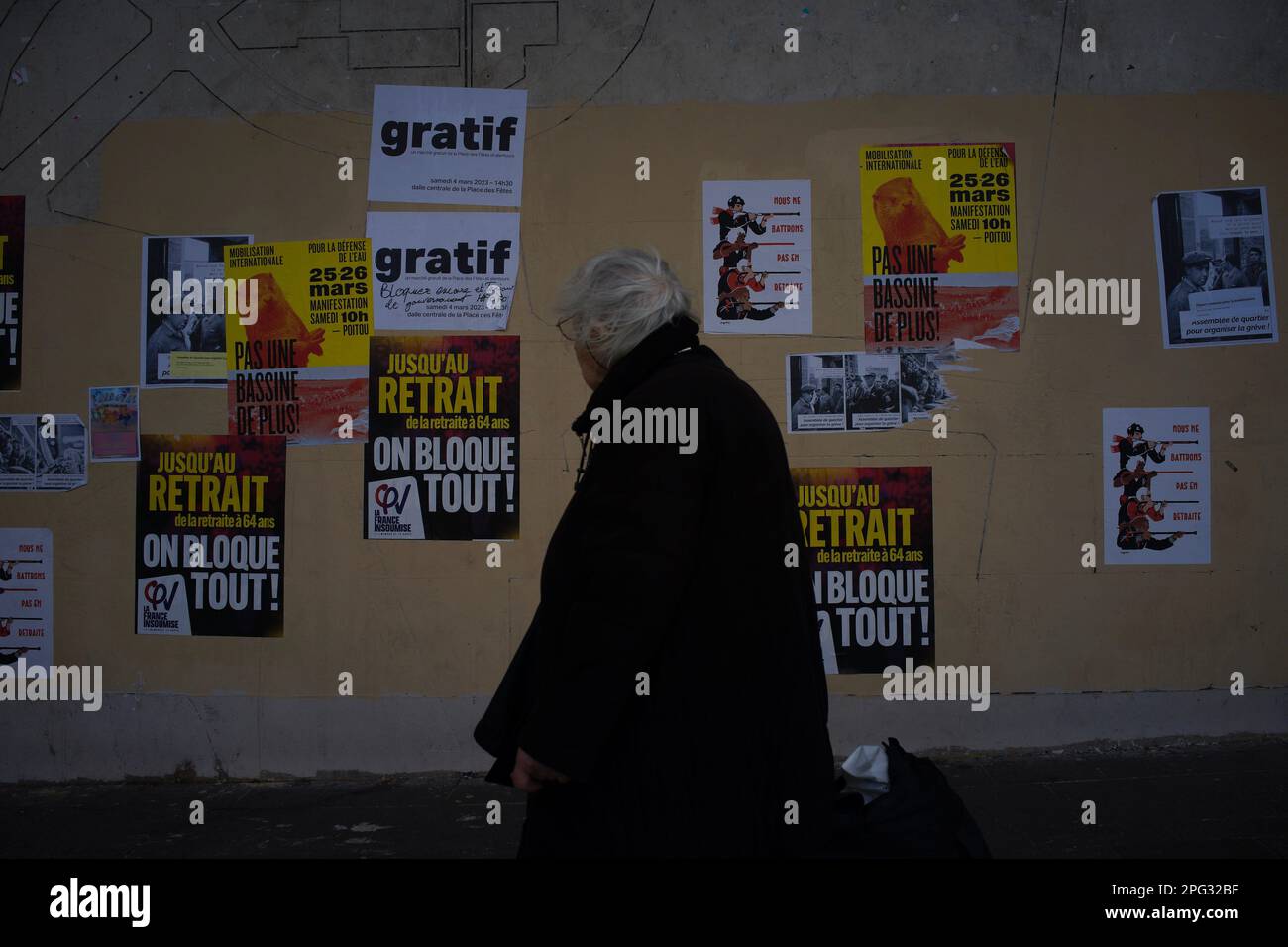 A pensioner walks past pension reform campaign posters as workers strike in opposition to Macron's controversial pension reforms, Place des Fêtes, 75019 Paris, France - Jane Burke, March 2023 Stock Photo