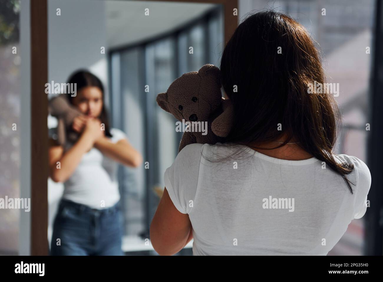 Young woman holds teddy bear and looks at herself into the mirror Stock Photo