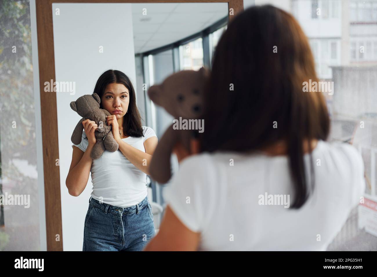 Young woman holds teddy bear and looks at herself into the mirror Stock Photo