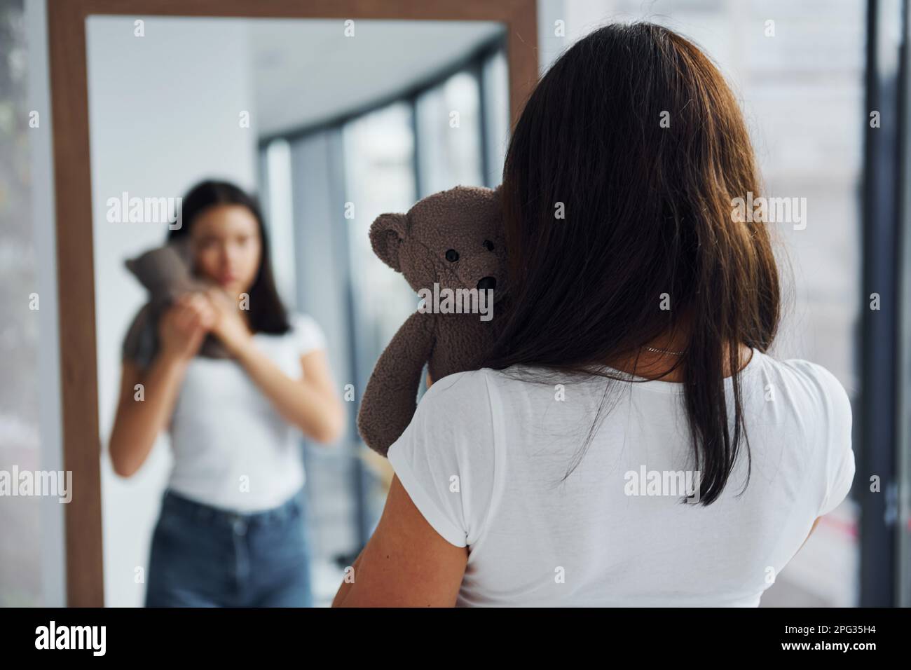 Young woman holds teddy bear and looks at herself into the mirror Stock Photo