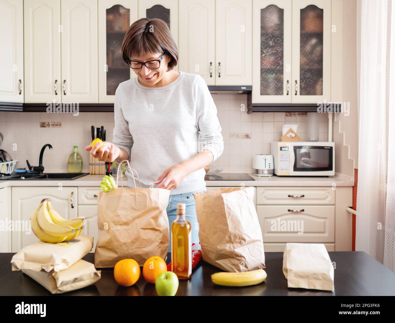 Woman sorts out purchases in the kitchen. Grocery delivery in paper bags. Subscription service from grocery store. Stock Photo