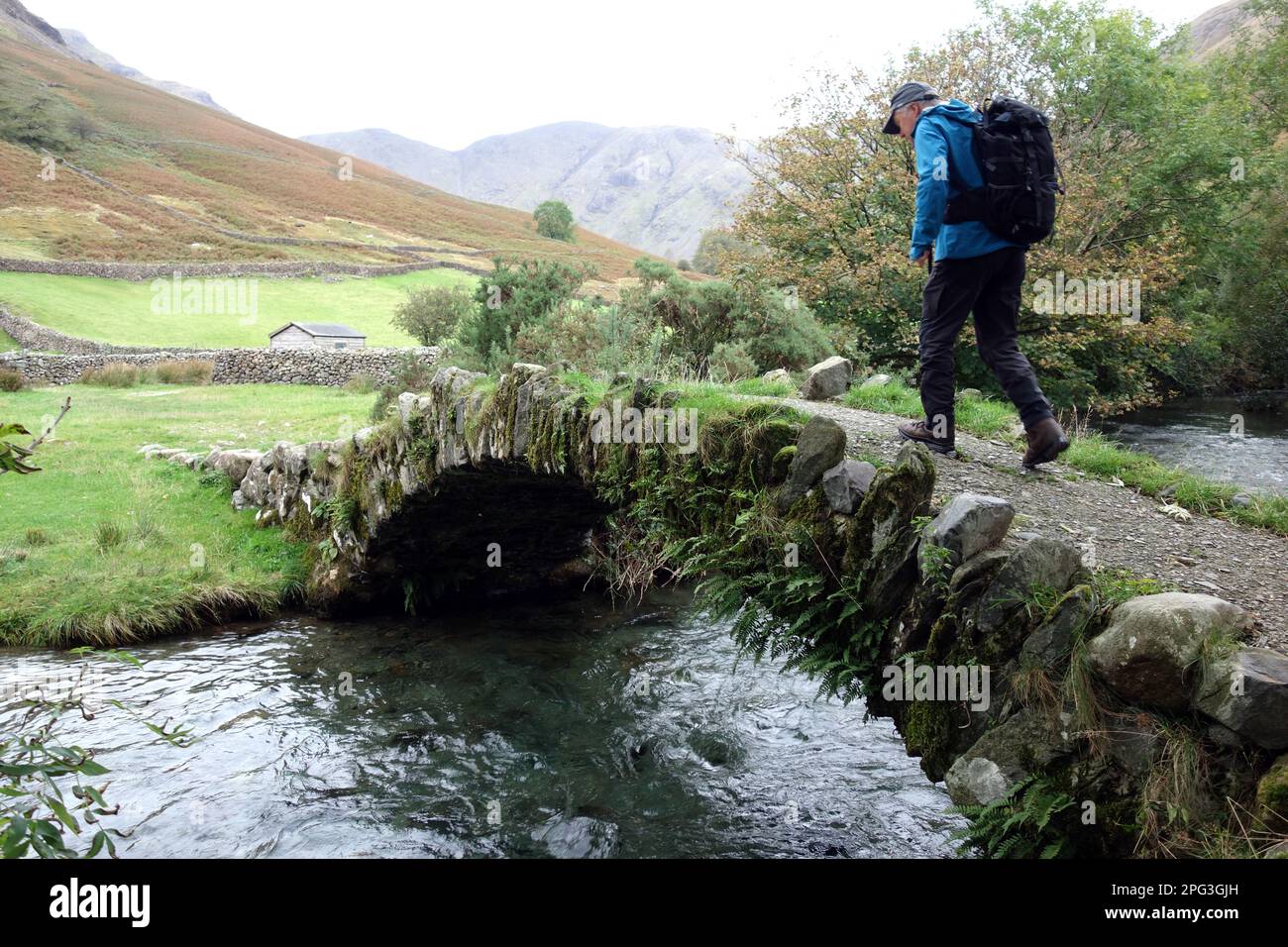 Man (Hiker) Walking over Single Stone Arched Packhorse Bridge over Mosedale Beck in Wasdale, Lake District National Park, Cumbria, England, UK. Stock Photo