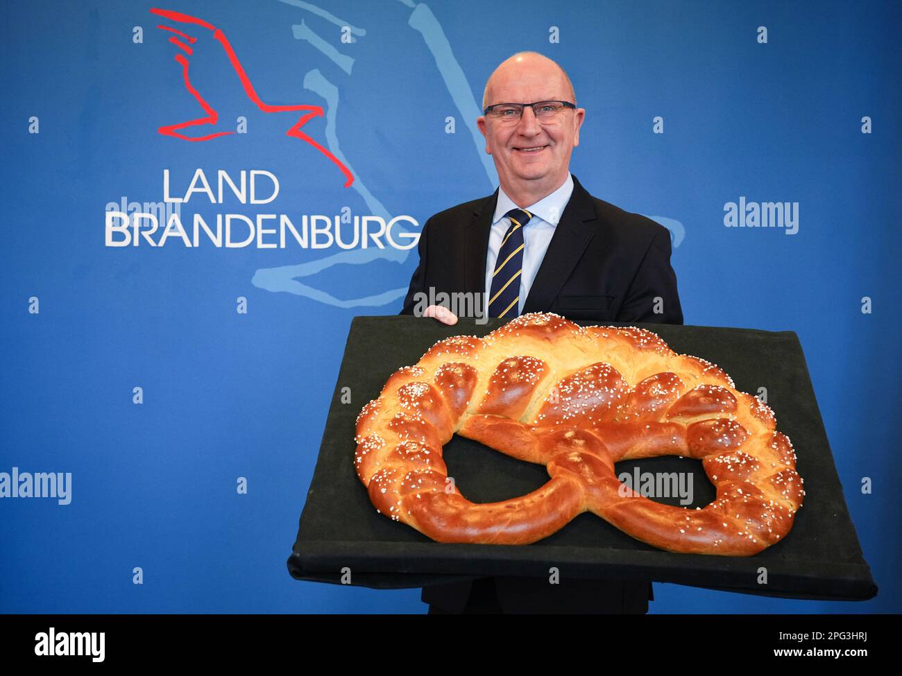 20 March 2023, Brandenburg, Potsdam: Brandenburg's Prime Minister Dietmar Woidke (SPD) symbolically receives a large pretzel made of baked dough for the 'Golden Pretzel' pin. The 'Golden Pretzel' is the seal of quality of the bakery and confectionery trade. Woidke received the award for his political services to the craft in Brandenburg. Photo: Soeren Stache/dpa Stock Photo