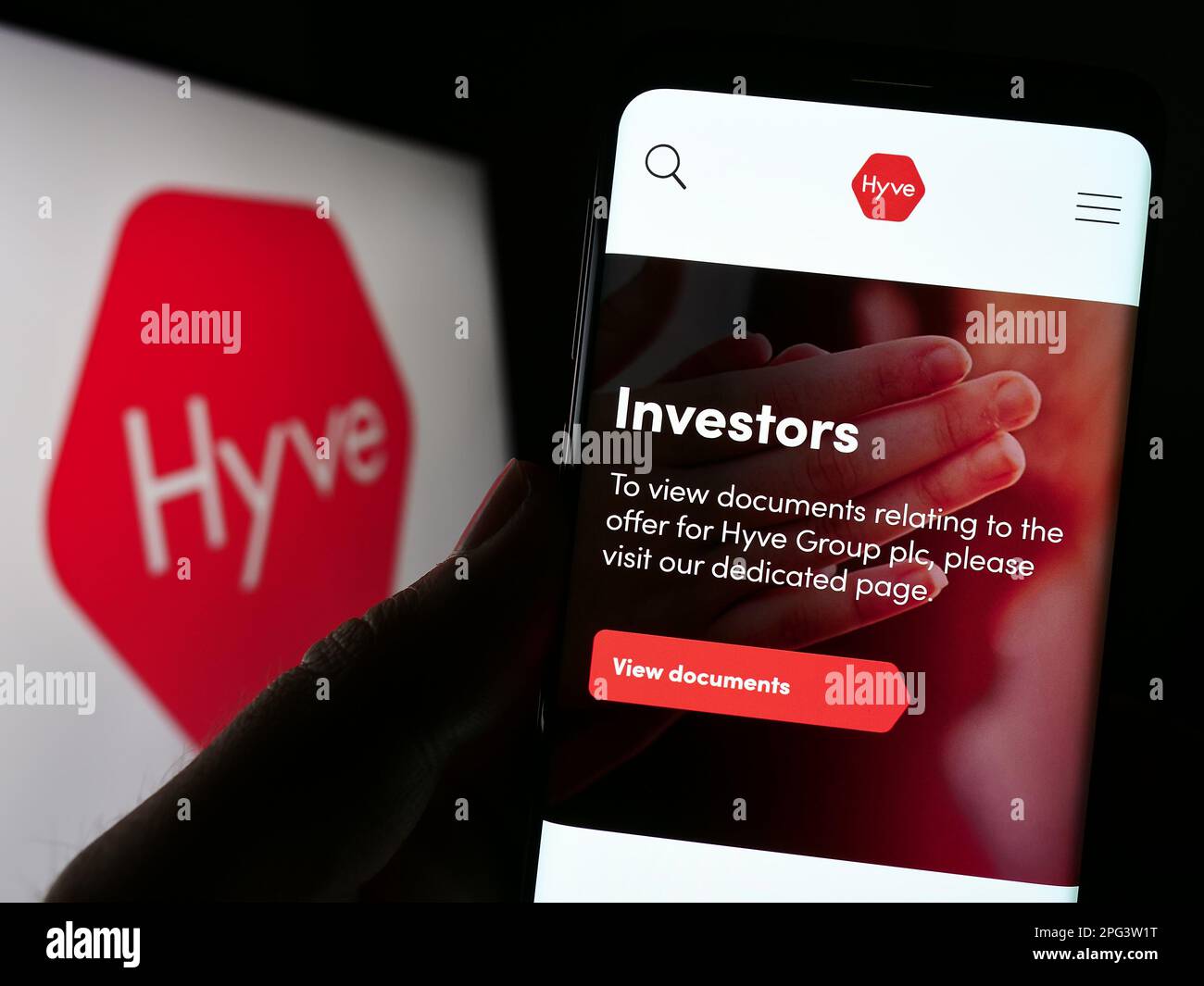 Person holding cellphone with webpage of British event management company Hyve Group plc on screen with logo. Focus on center of phone display. Stock Photo