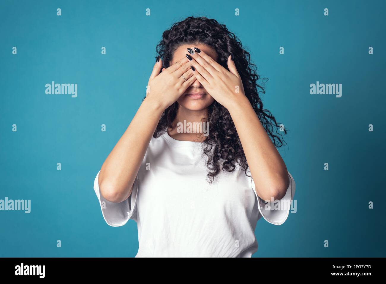 Shot of serious confident curly woman, wears casual basic solid white t-shirt, covering her face isolated over blue background. Expressions Stock Photo