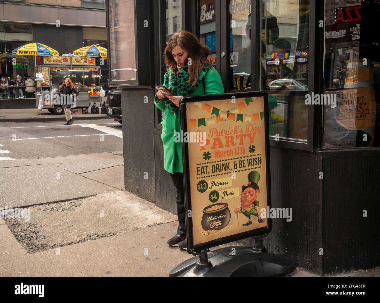 Distracted woman in her St. Patrick’s Day green coat in front of a bar advertising its St. Patrick’s Day party, in Midtown Manhattan on Sunday, March 12, 2023. (© Richard B. Levine) Stock Photo