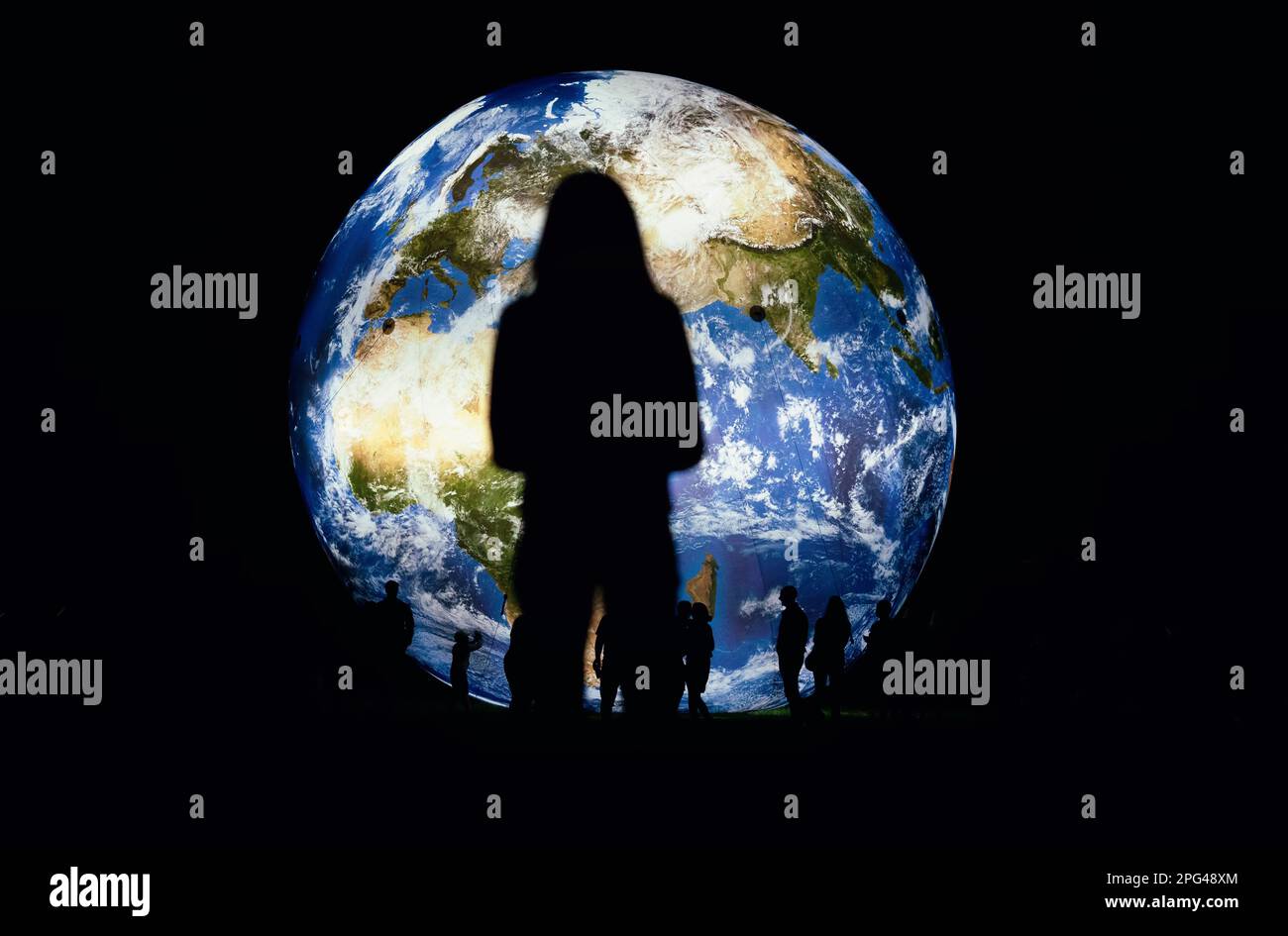 Silhouettes of people and young woman standing in front of large inflatable model of planet Earth glowing at night, view from behind Stock Photo