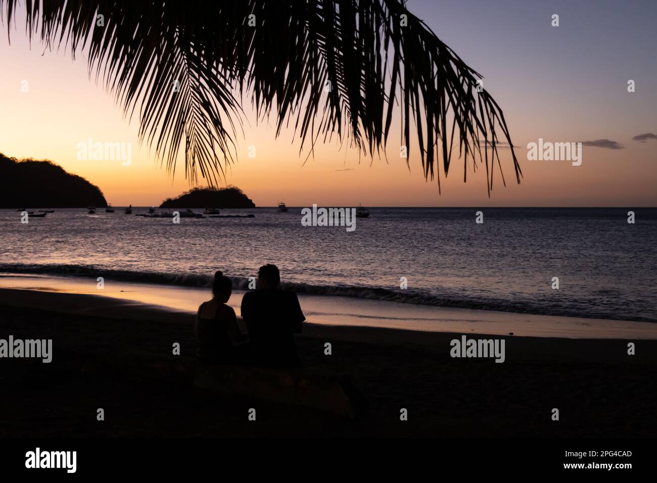 A beautiful sunset in Costa Rica with a silhouette of a man and woman sitting on the beach. A palm branch hangs above their heads. Stock Photo