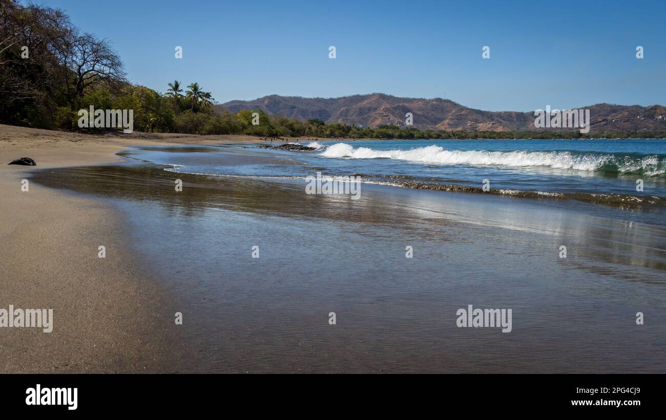 A peaceful tropical image of waves crashing onto a smooth sand beach in Costa Rica. In the distance are hills and palm trees. Stock Photo