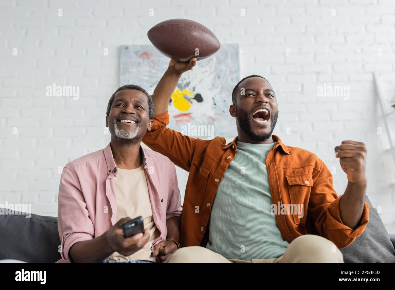 Excited african american man holding rugby ball near dad with remote controller in living room,stock image Stock Photo