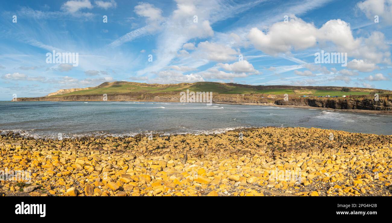 Orange clay rocks in foreground looking out to Kimmeridge bay and Jurassic cliffs and carpark on a sunny day with light clouds Stock Photo