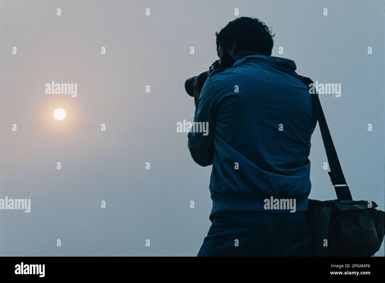 photographer eyeing up a shot is silhouetted by a foggy sky and the hazy ball of the sun Stock Photo