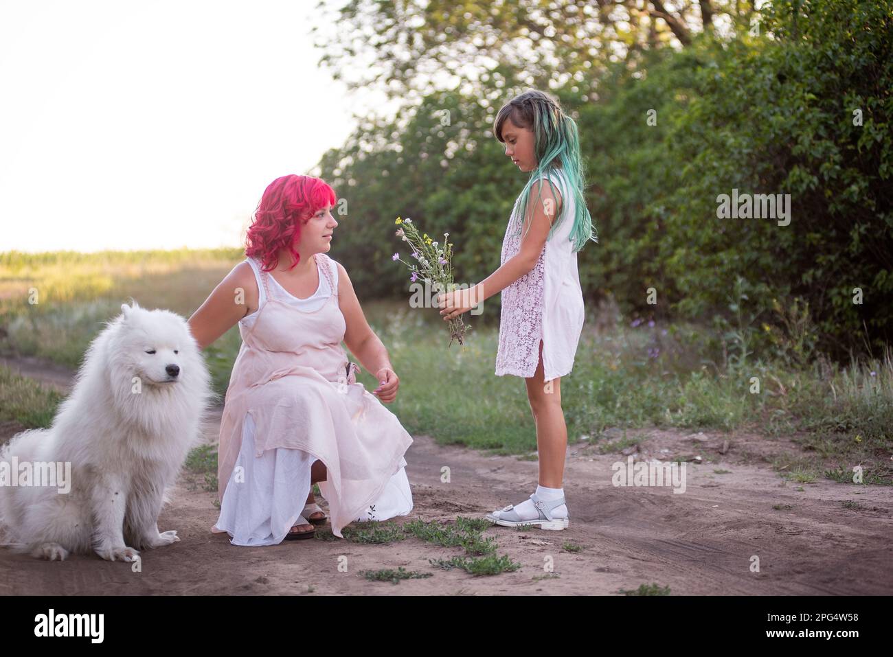 Girl with green hair gives wild flowers to mother with pink. Traveling with Samoyed dog, pets to nature. The tenderness of motherhood, one parent. Div Stock Photo