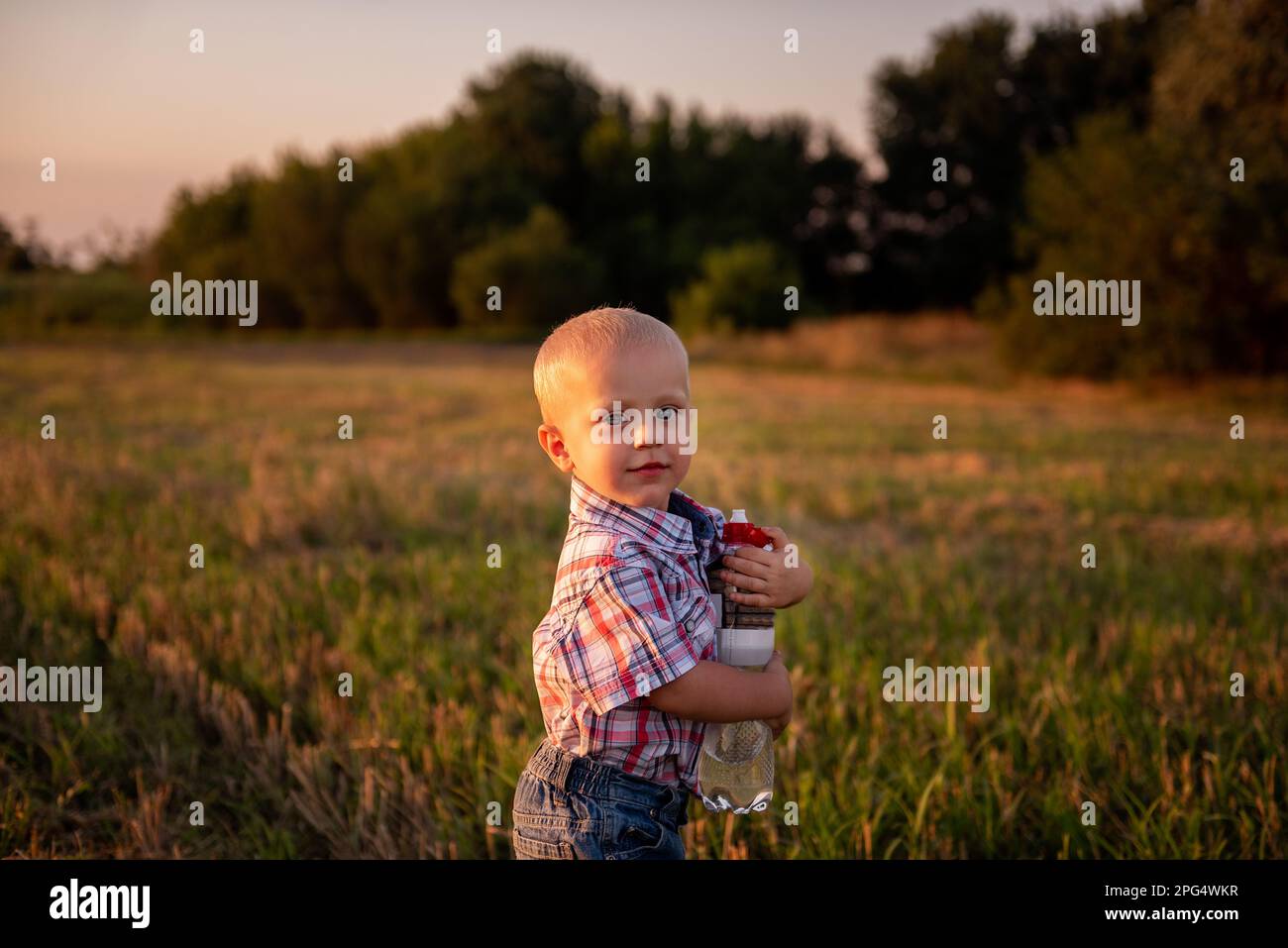 Little boy in plaid shirt holds bottle of water in hands, in mowing field of wheat in rays of the sunset. Carefree childhood in rural areas. Concept o Stock Photo