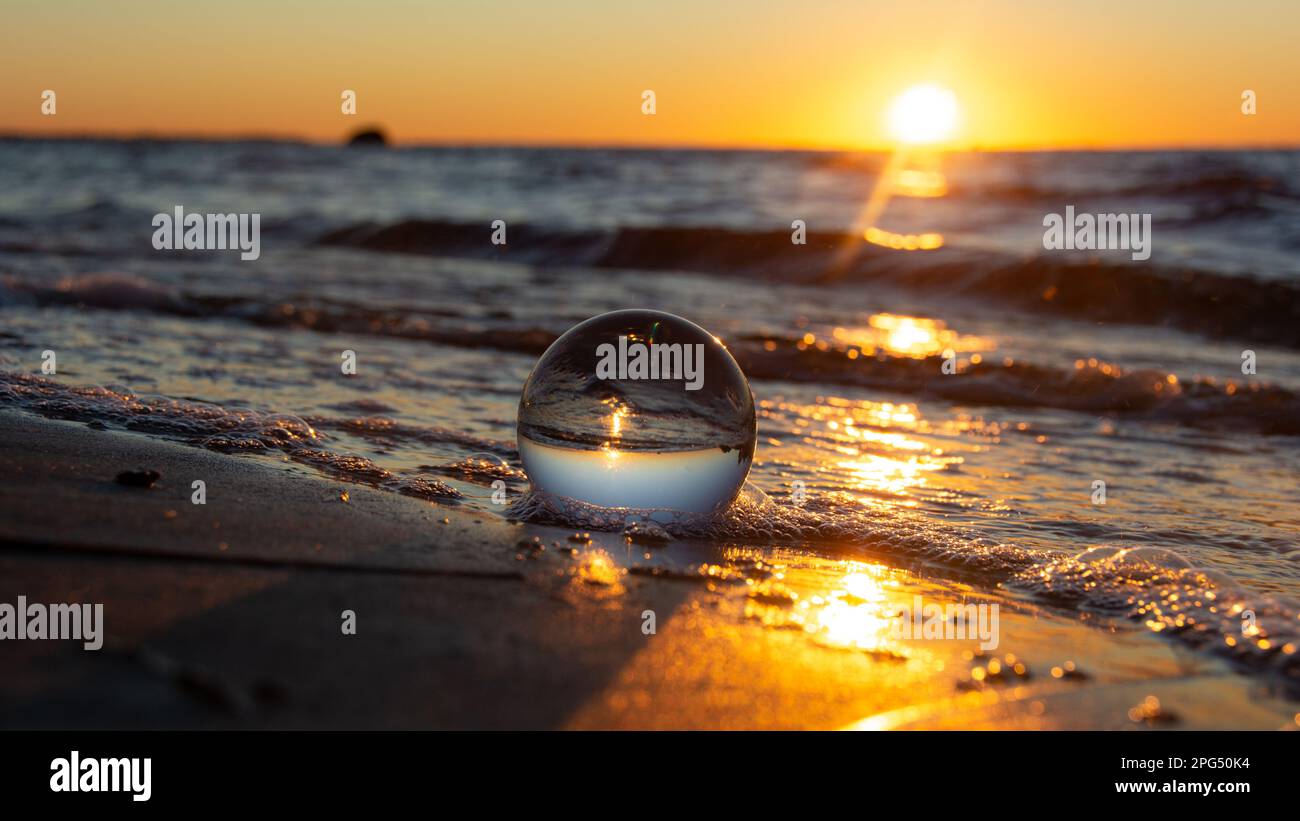A glass ball lies in the waves on the sandy beach, the sea and the setting sun are reflected in the ball Stock Photo