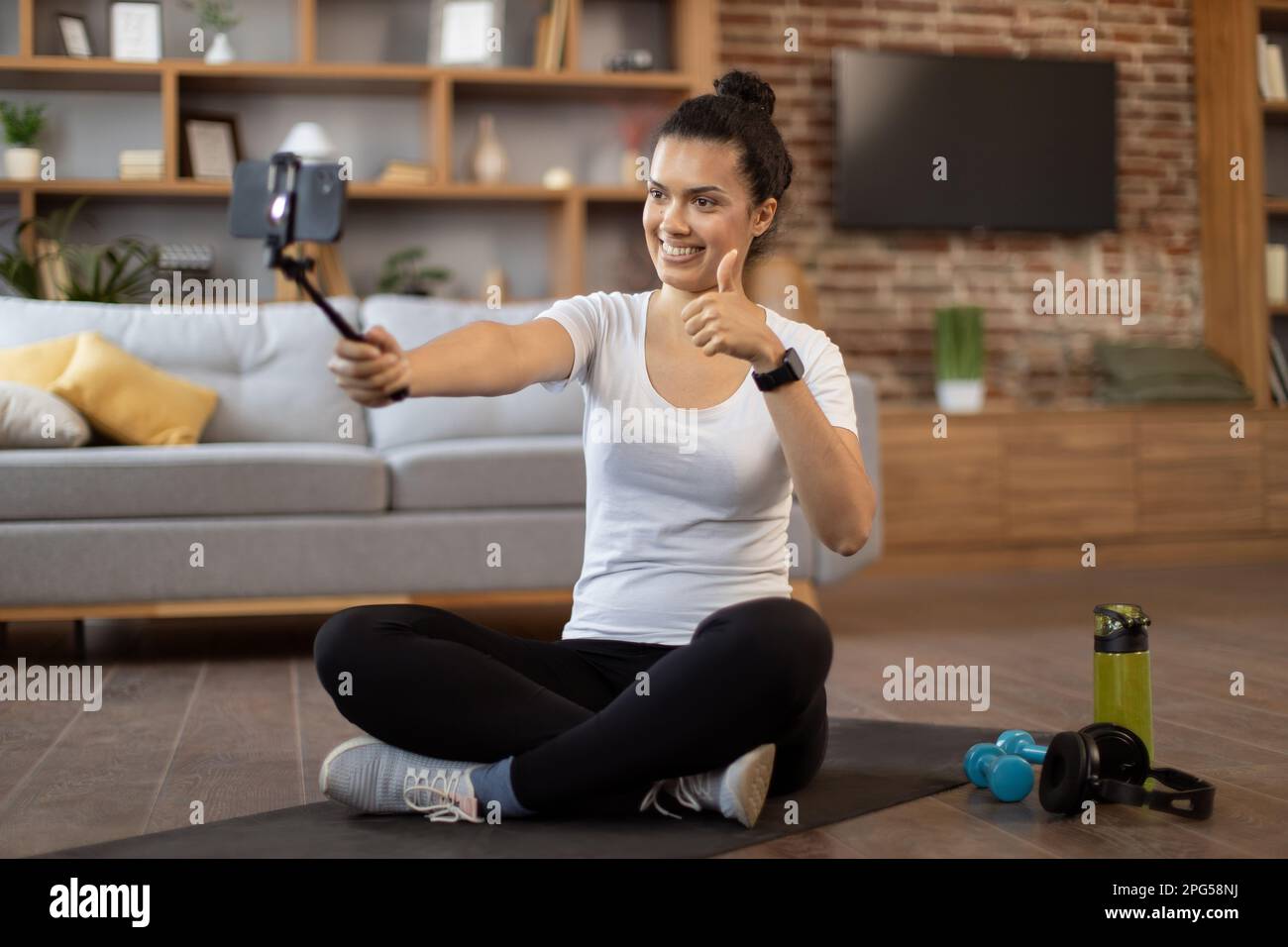 Young active multiracial woman in sportswear using cell phone on tripod while live streaming fitness tutorial for sports vlog indoors