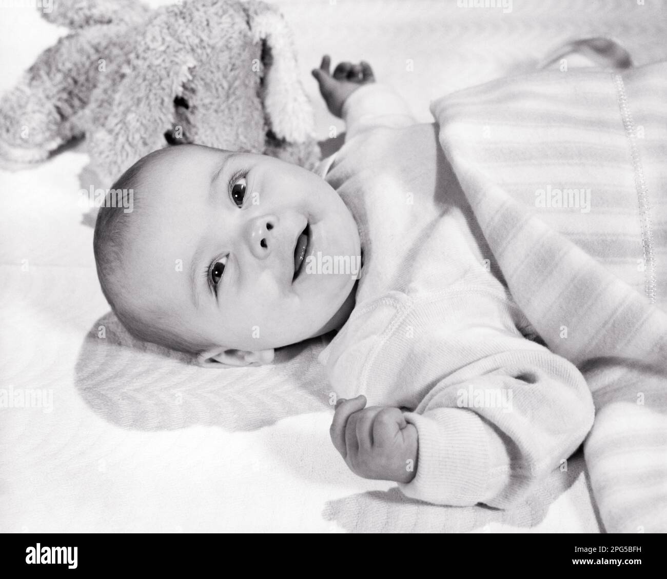 1960s SMILING BABY LYING DOWN WITH TOY AND BLANKET LOOKING UP AT CAMERA - b21396 HAR001 HARS CHEERFUL HIGH ANGLE AND EXCITEMENT UP SMILES CONNECTION JOYFUL PLEASANT AGREEABLE CHARMING GROWTH JUVENILES LOVABLE PLEASING ADORABLE APPEALING BABY GIRL BLACK AND WHITE CAUCASIAN ETHNICITY HAR001 OLD FASHIONED Stock Photo