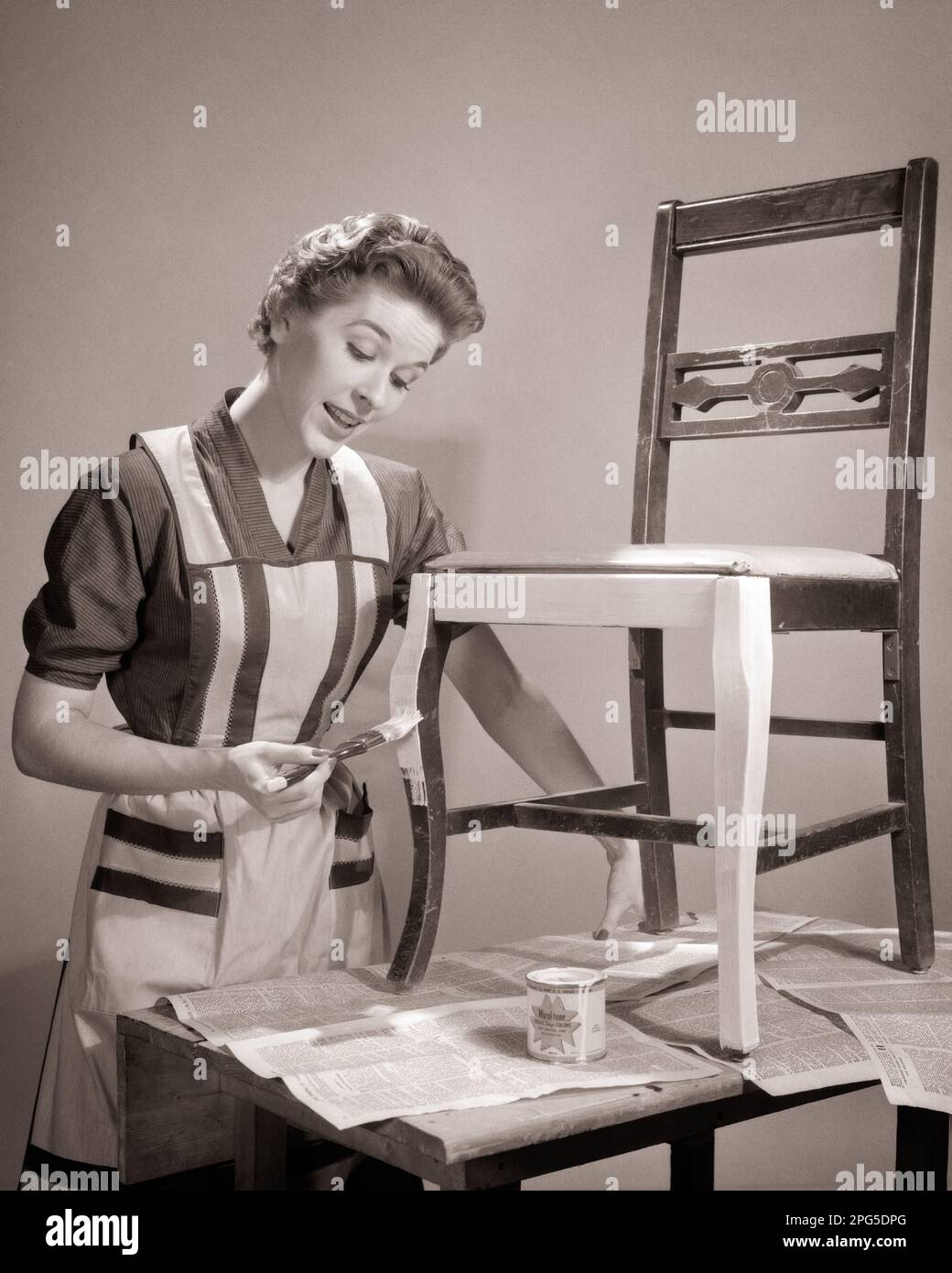 1950s HOUSEWIFE WEARING APRON PAINTING DARK WOOD DINING ROOM CHAIR LIGHTER COLOR ON TABLE COVERED WITH NEWSPAPER FOR PROTECTION  - p1942 DEB001 HARS PERSONS INSPIRATION CARING CONFIDENCE B&W HOMEMAKER COVERED HOMEMAKERS CHORE LEISURE PROTECTION HOUSEWIVES PAINTED TASKS DEB001 LIGHTER PRIMER CREATIVITY MID-ADULT MID-ADULT WOMAN PAINT CAN SOLUTIONS TASK BLACK AND WHITE CAUCASIAN ETHNICITY OLD FASHIONED Stock Photo