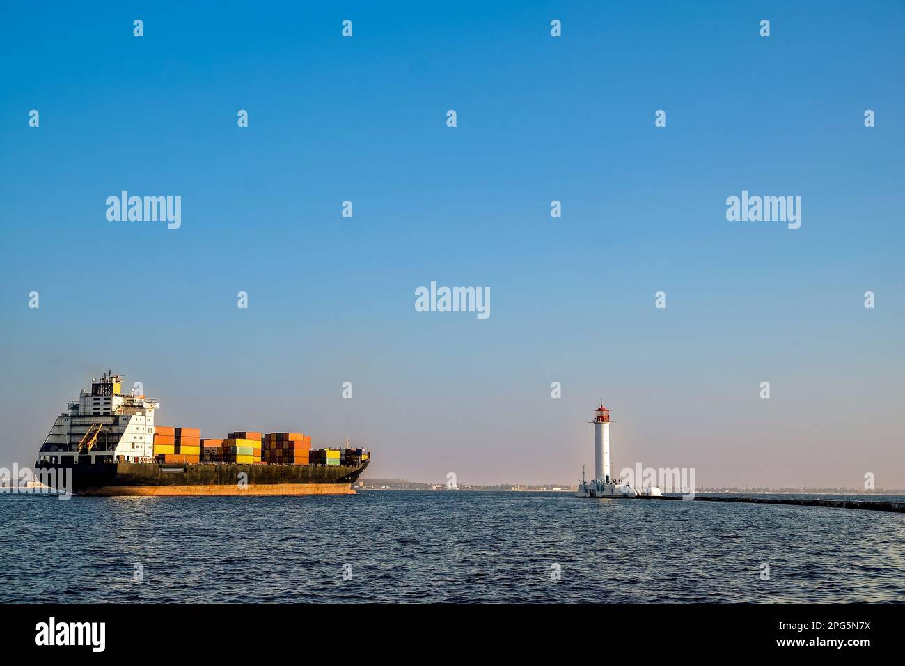 A container ship leaves the port near the lighthouse. Evening Stock Photo