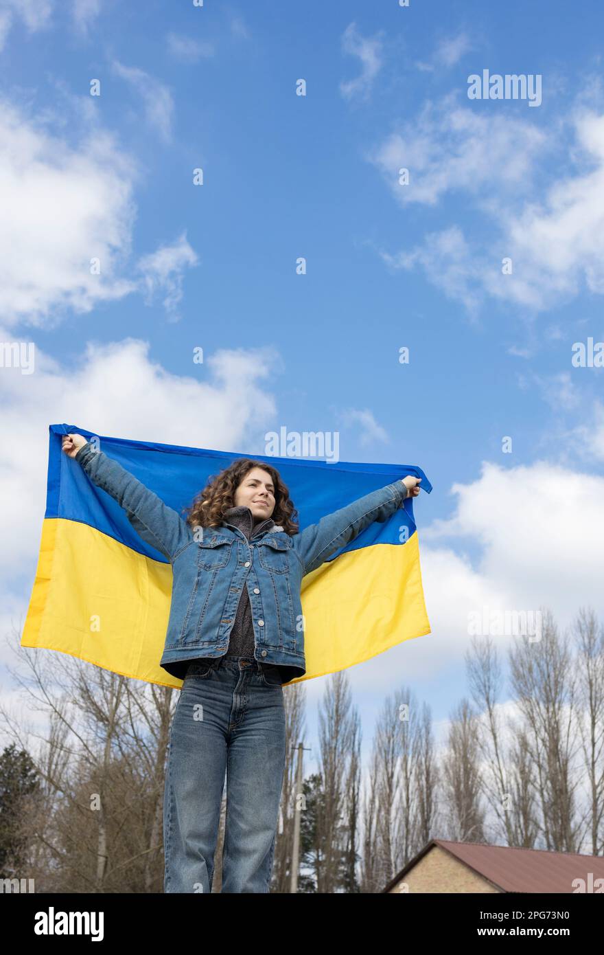 young woman, an adult girl, holds a Ukrainian yellow-blue flag behind her back against the sky. Ukrainians are against the war. Drawing attention to m Stock Photo