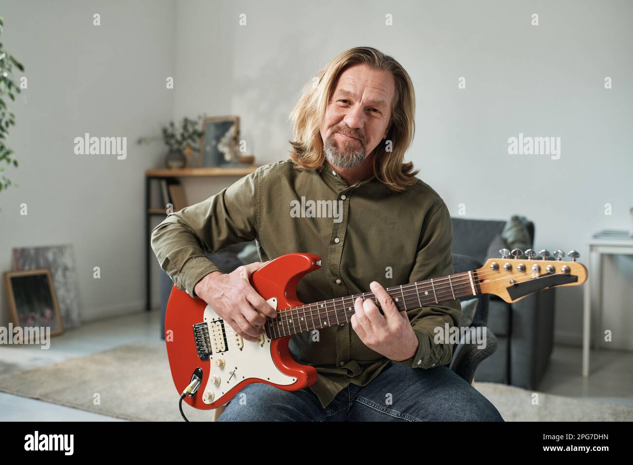 Portrait of mature guitarist with long hair looking at camera while playing guitar online Stock Photo