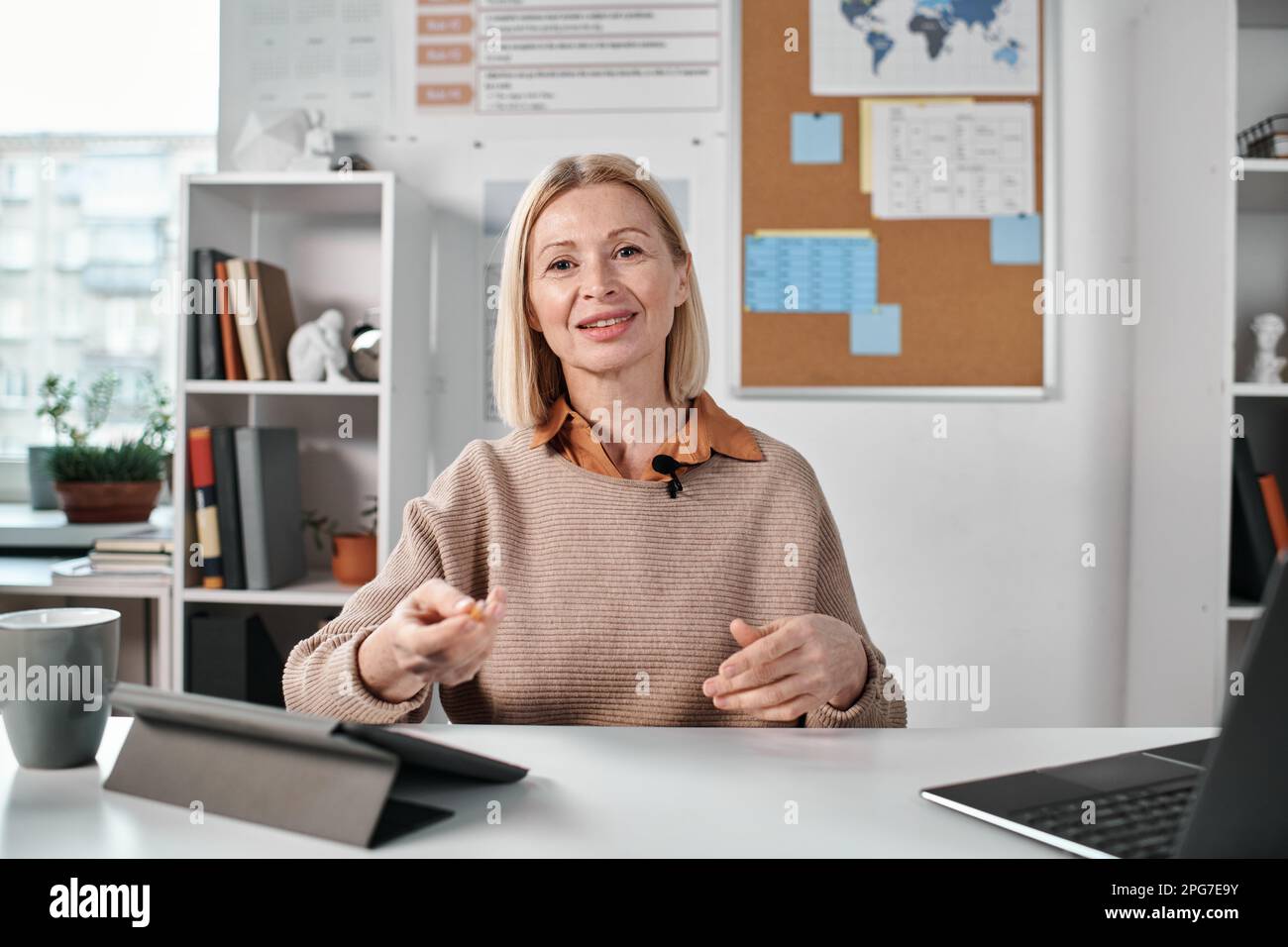 Portrait of mature teacher looking at camera while sitting at table in class and giving online lesson to students Stock Photo