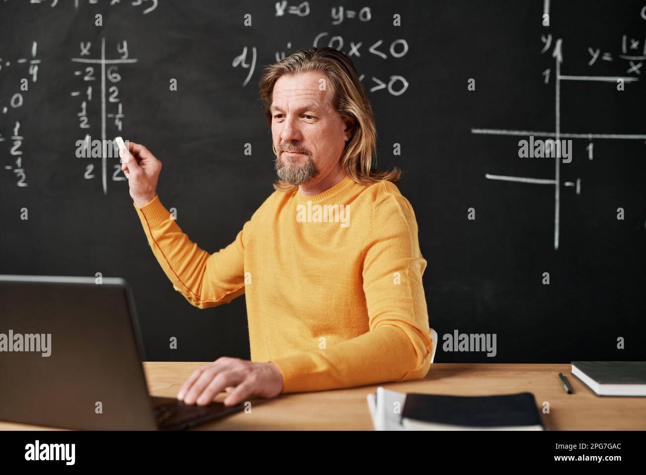 Mature bearded teacher writing on blackboard and using laptop for online lesson in the classroom Stock Photo