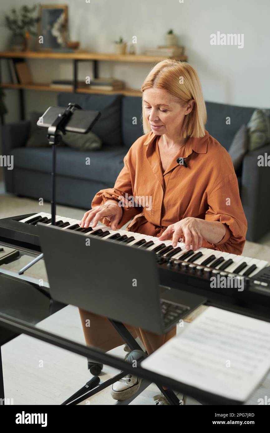 Mature teacher sitting at table in front of laptop and playing piano online for her student Stock Photo