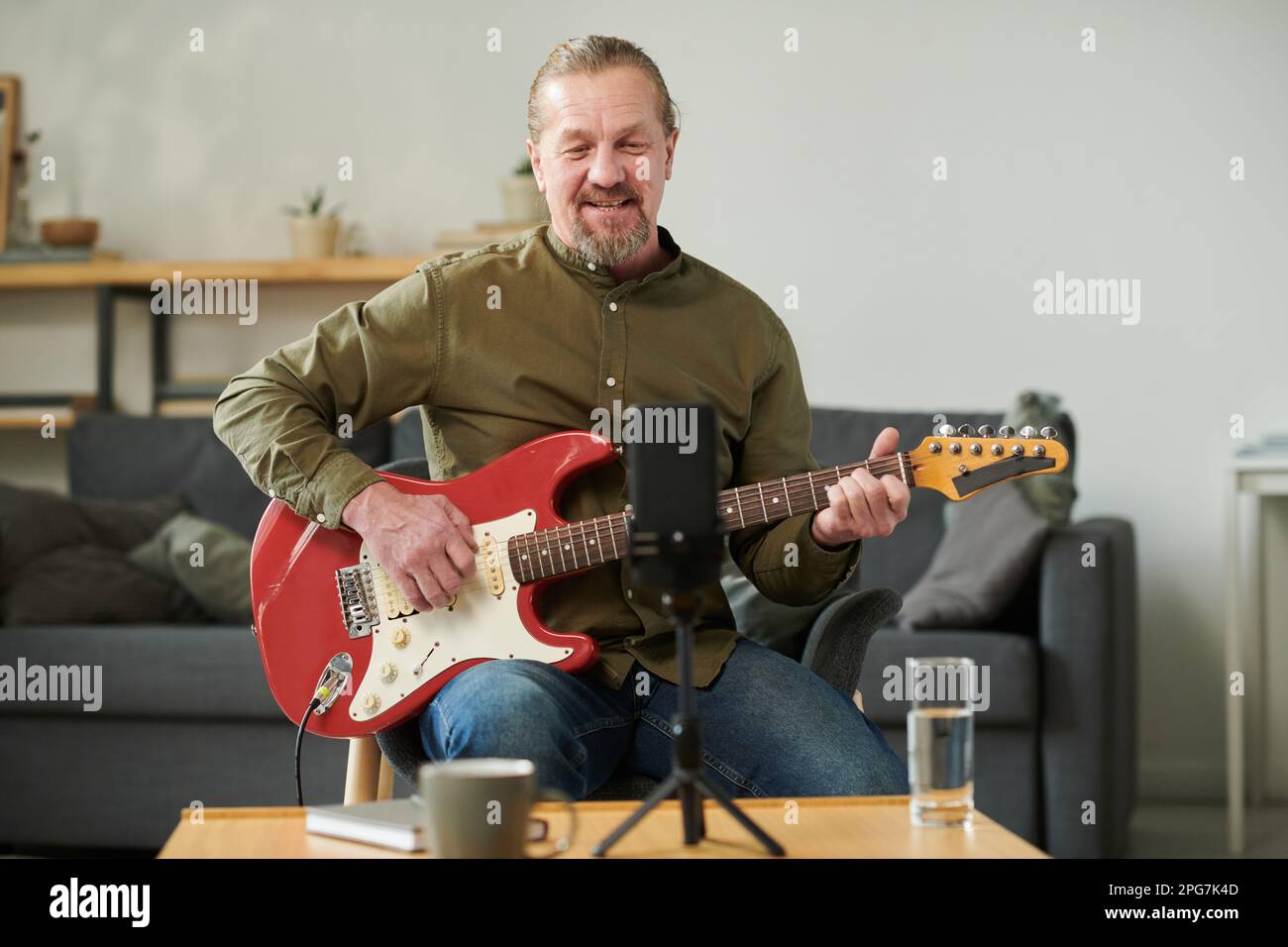 Mature teacher sitting in front of smartphone and playing guitar during online lesson with student Stock Photo