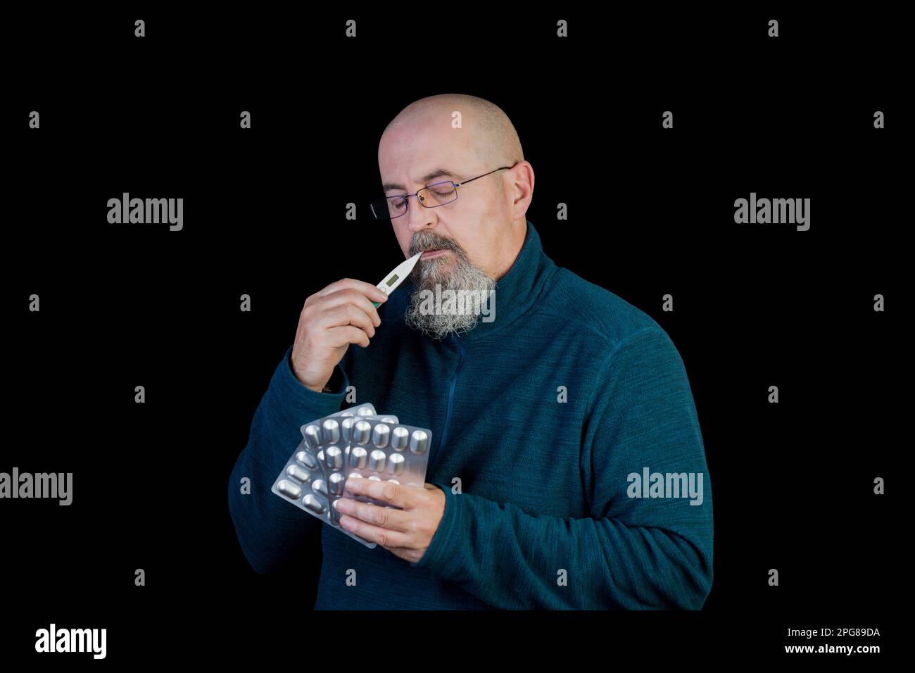 A middle-aged man checking his temperature with a bunch of pills in his hands, isolated on black background Stock Photo