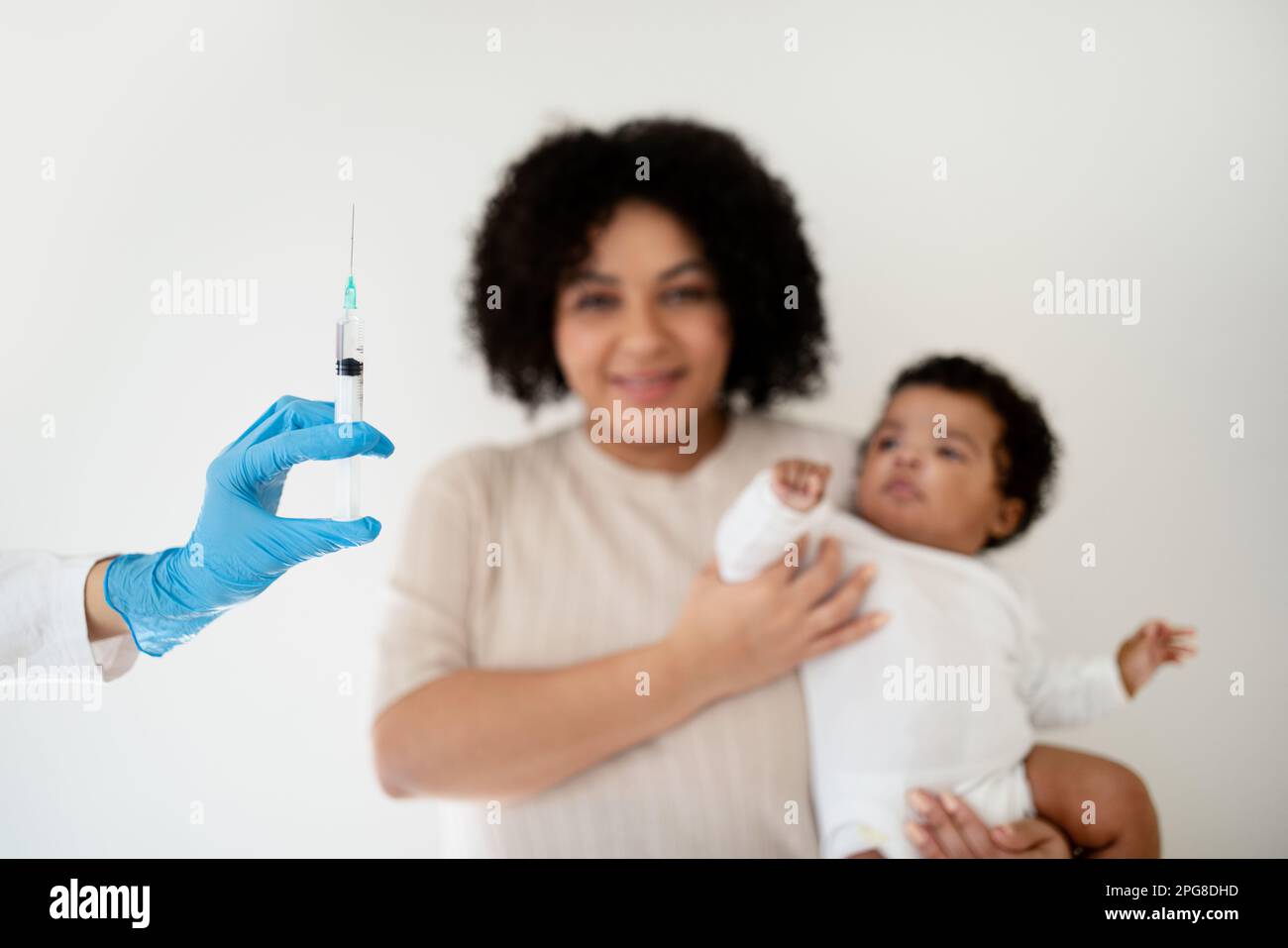 Smiling loving young african american woman holding baby in her arms, looks at doctor hand with syringe Stock Photo