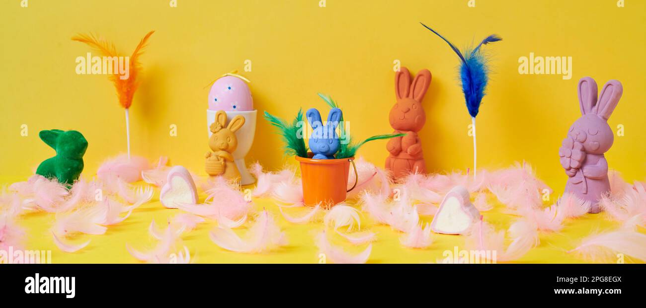 some easter bunnies of different colors, a pink easter egg in a white eggcup and some pink feathers  on a yellow background, in a panoramic format to Stock Photo