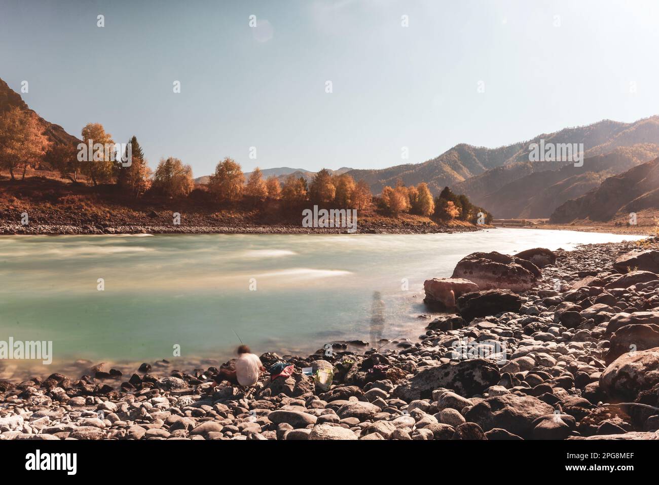 Blurred silhouettes of two women resting with a fishing rod on the bank of the mountain river Katun between the rocks during the day. Long exposure. Stock Photo