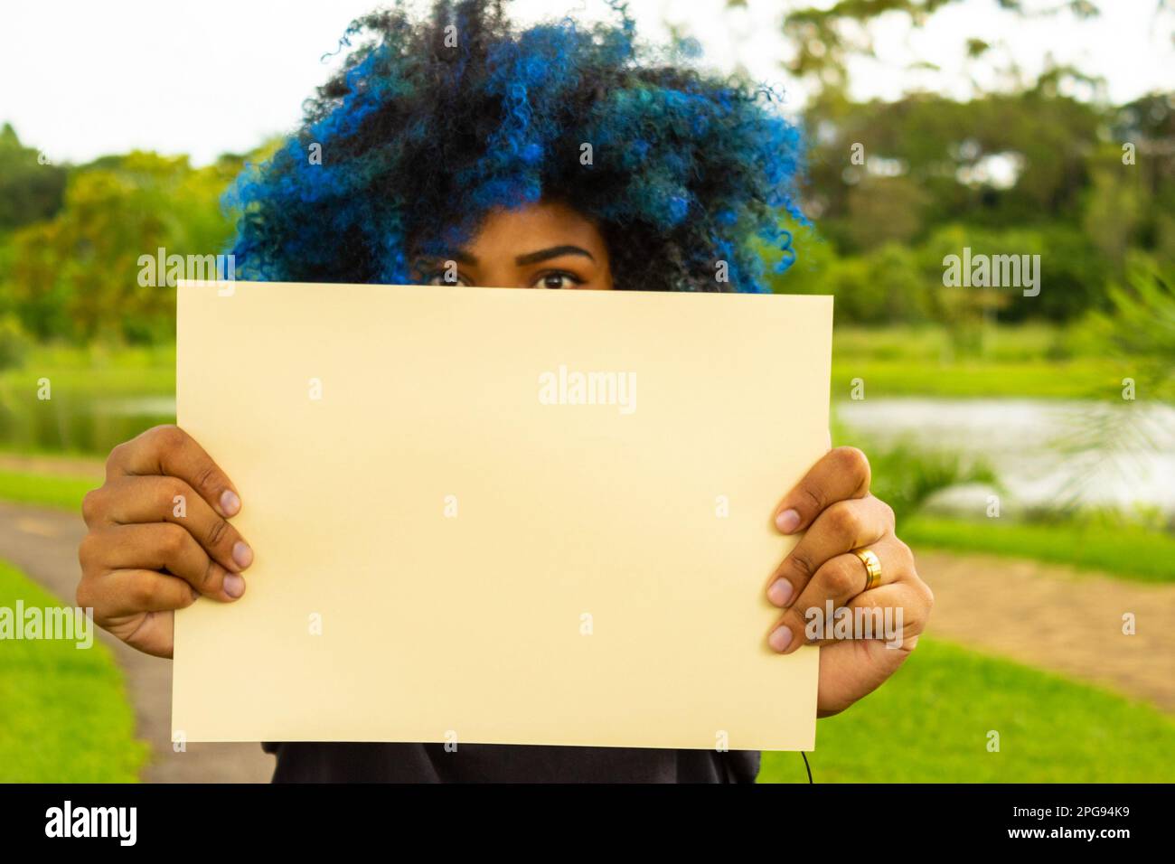 Goiania, Goias, Brazil – March 21, 2023: A young woman, with hair dyed blue, her face hidden behind a blank poster, with a landscape in the background Stock Photo