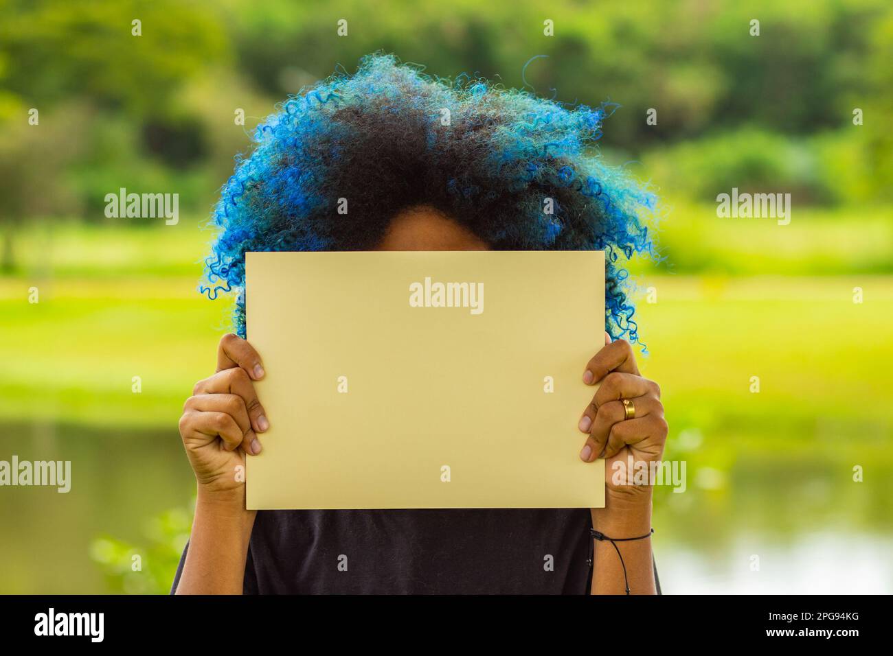 Goiania, Goias, Brazil – March 21, 2023: A young woman, with hair dyed blue, her face hidden behind a blank poster, with a landscape in the background Stock Photo