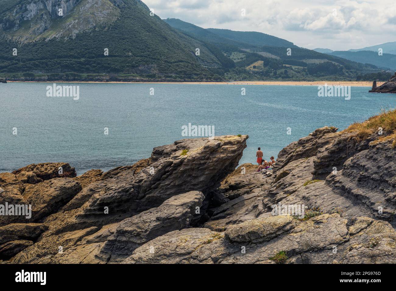 two young women and a man sunbathing in swimsuits on the rocks of the town of Sonabia, the Cantabrian Sea and the mountains in the background, Spa Stock Photo