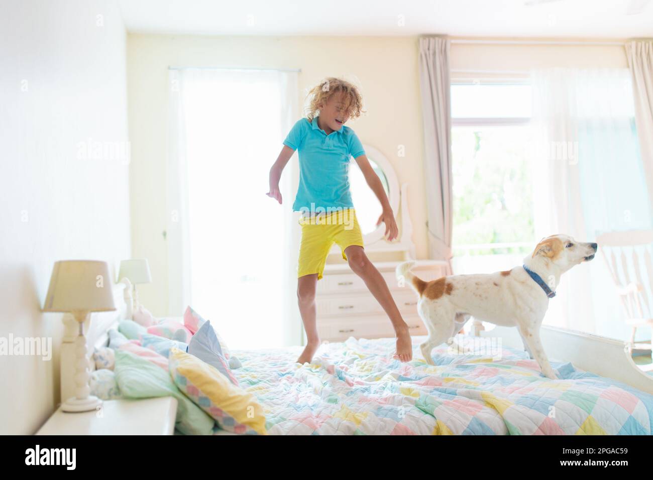 Child jumping on bed in white bedroom with big window. Kids play at