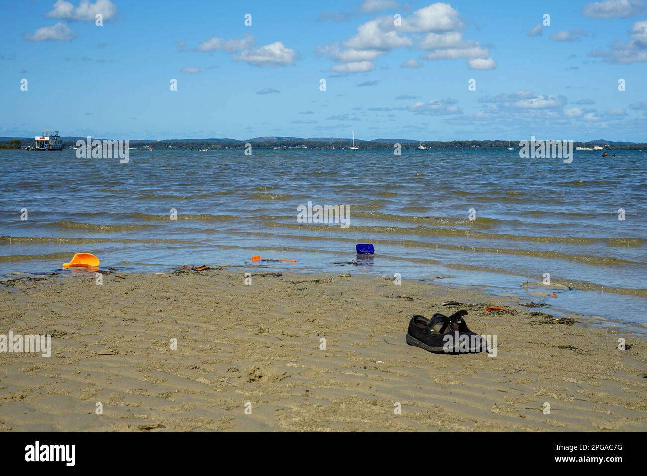 Black shoes, and colourful plastic Beach toys by the waters edge at Thompsons Beach, Victoria Point Queensland. Macleay Island in the distance. Stock Photo
