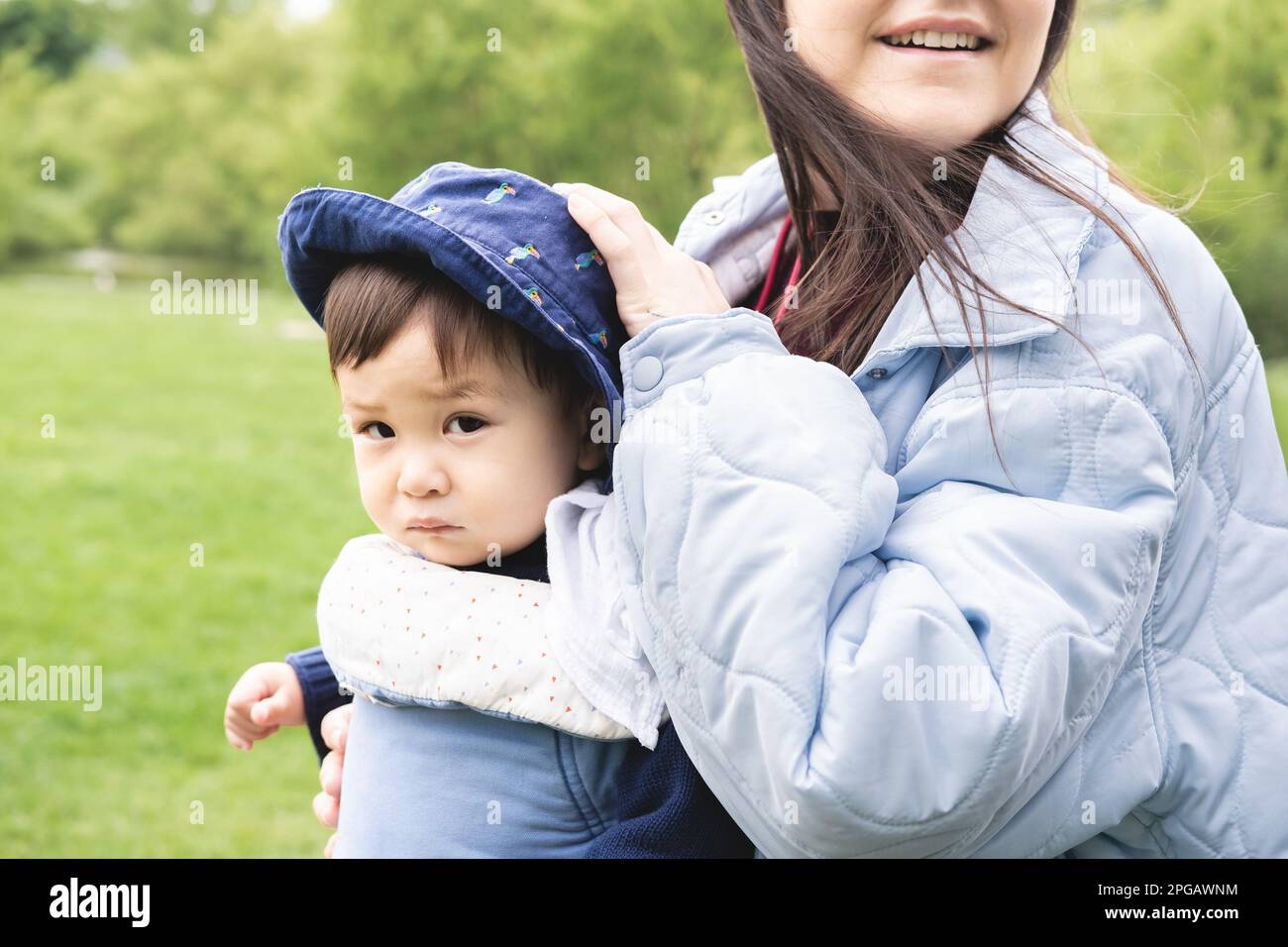 Close-up photo of a male infant carried by his smiley mother on a baby carrier wearing a blue hat walking at the park. He is feeling poor and looking Stock Photo