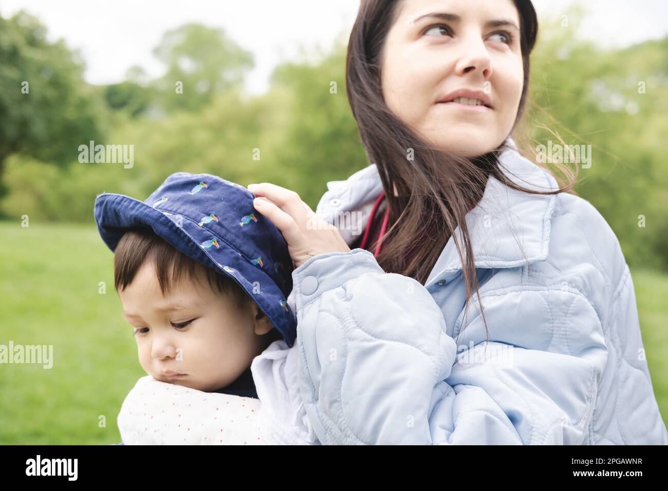 Close-up photo of a male infant carried by his smiley mother on a baby carrier wearing a blue hat walking at the park. He is feeling poor and ill. Mul Stock Photo