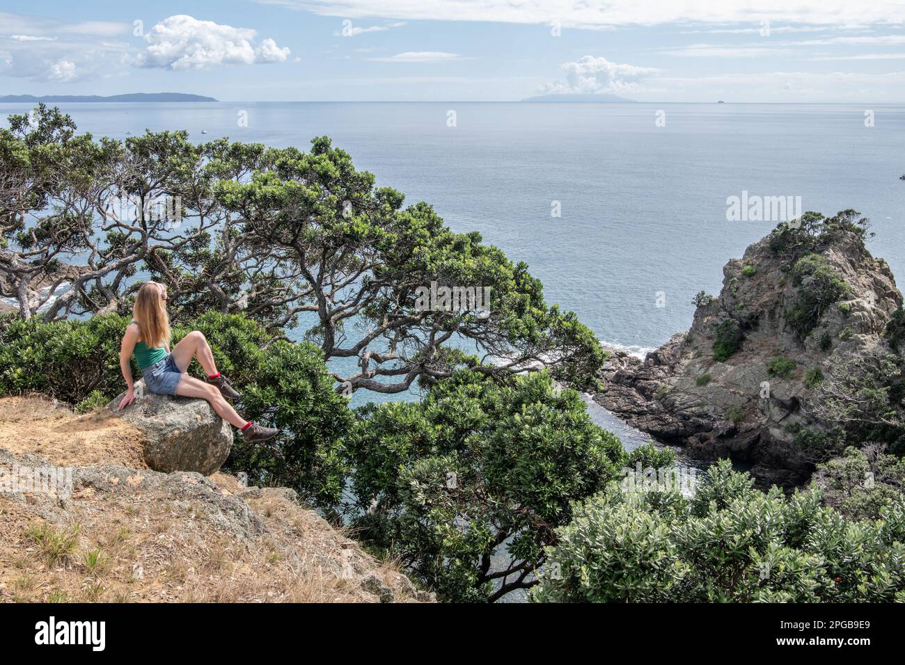 A woman sits on the rocky coast of Tiritiri Matangi island in the Hauraki gulf looking over the pacific ocean with little barrier island visible. Stock Photo