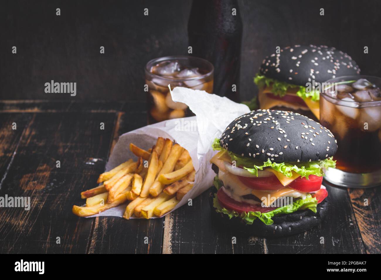Set of black burgers with meat patty, cheese, tomatoes, mayonnaise, french fries in a paper cup and glass of cold cola soda with ice. Dark wooden Stock Photo