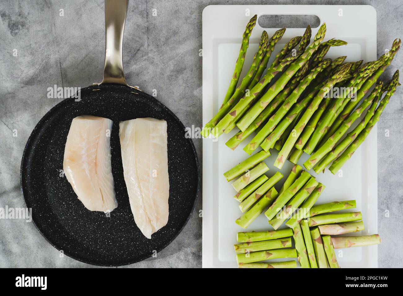 Raw cod fillet on a black frying pan, and raw asparagus on a white cutting board close-up on the kitchen table, flat lay Stock Photo