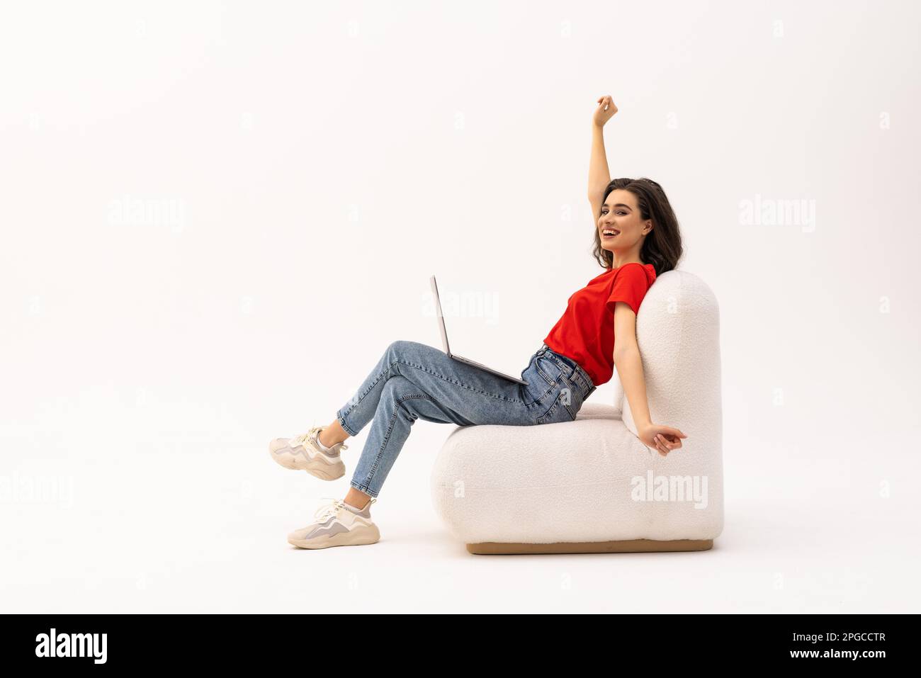 Euphoric young woman celebrating success with laptop, raising arms up, sitting in armchair on white background. Joyful millennial lady with portable c Stock Photo