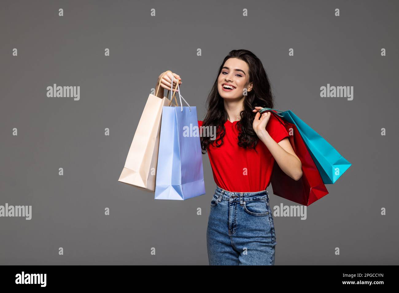 Photo of excited adorable woman with long curly hair smiling and carrying colorful shopping bags isolated over gray background Stock Photo