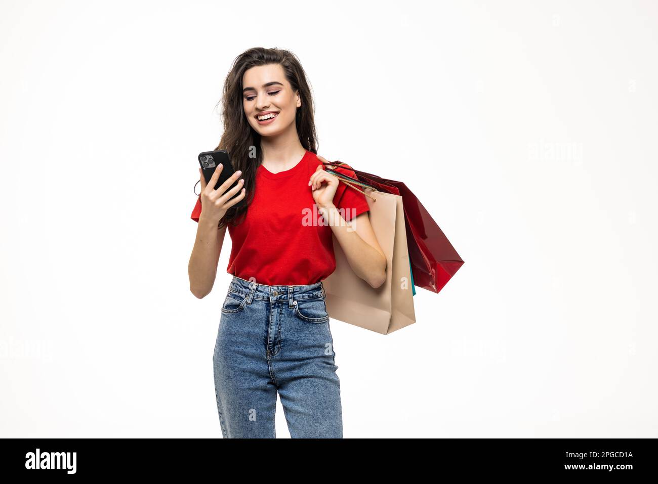 Online shopping. Beautiful young woman holding shopping bags and using her smartphone with smile while standing against white background Stock Photo