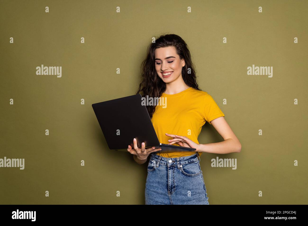 Smiling happy brunette woman in casual white shirt and jeans is working on laptop on khaki background and looking at camera. Remote, distance work, jo Stock Photo